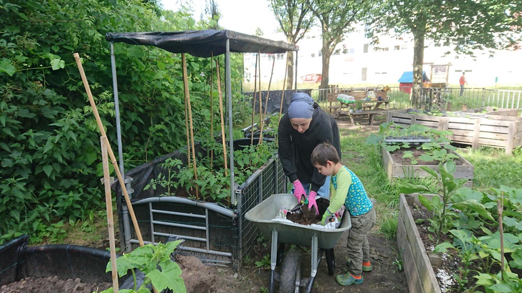 Eine Frau mit Kopftuch und ein Kind arbeiten zusammen im Geimeinschaftsgarten. Sie nehmen aus einer Schubkarre Erde.