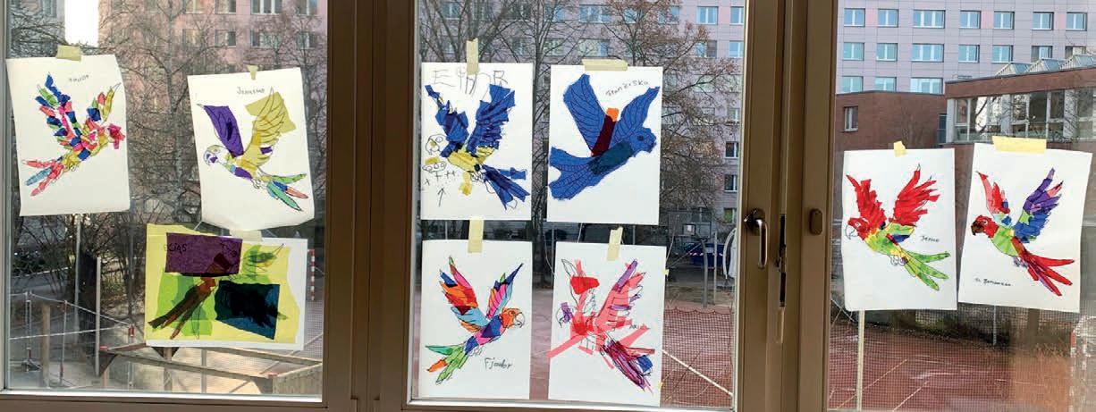 An der Fensterfront des Klassenzimmers viele verschiedene bunte Papagaien-Bilder aufgehängt. Im Hintergrund sieht man den Sportplatz und einen Plattenbau.