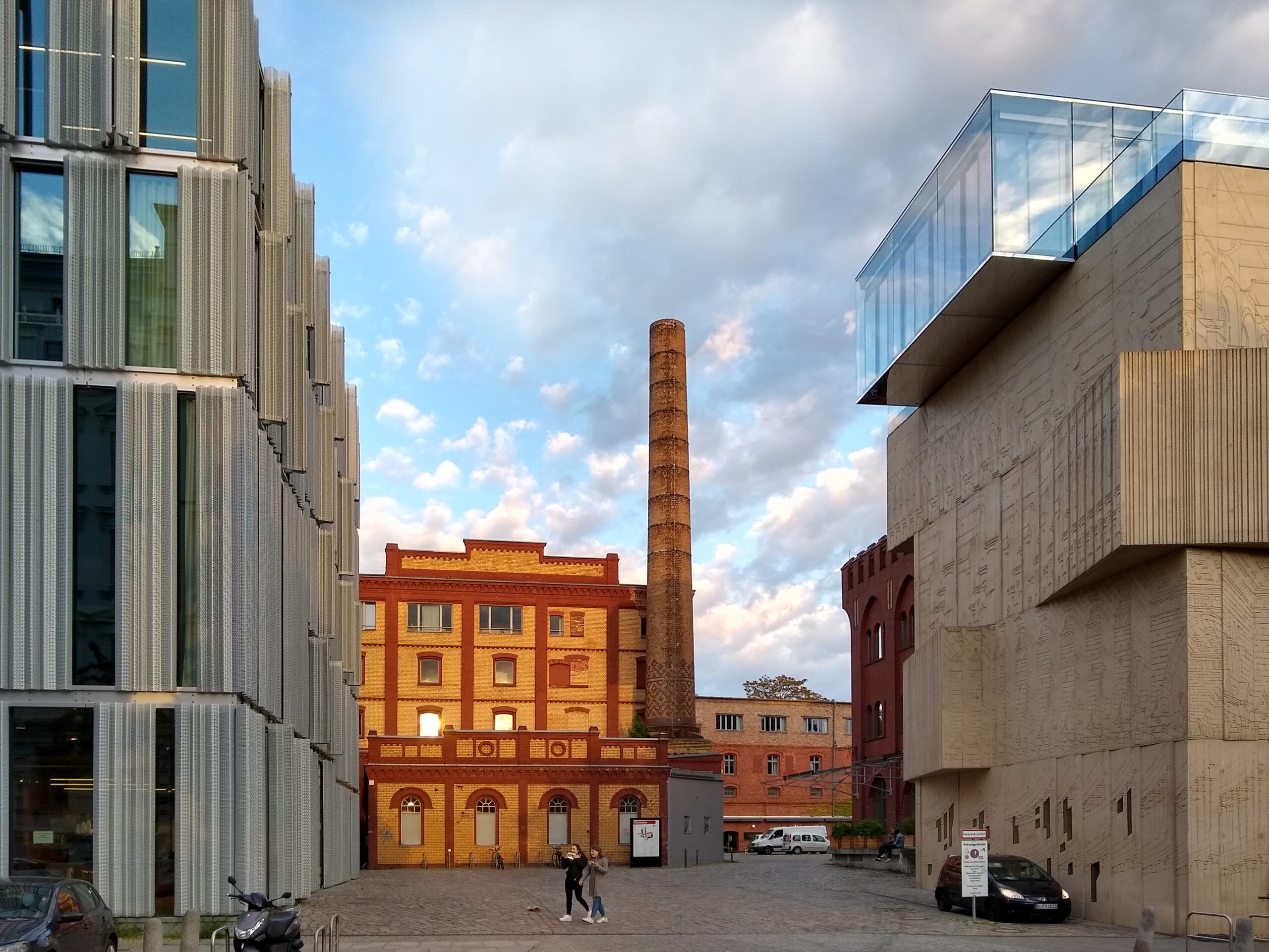 Am Eingang zum Christinenstraße zum Pfefferberg laufen in der Abendsonne vor dem historischen Fabrikgebäude mit gelb und roten Backsteinen zwei Personen über den Platz. Rechts ist das moderne Gebäude des Museums für Architekturzeichnung aus Beton zu sehen und links das ebenfalls neu errichtete Atelierhaus mit seiner Fensterfront mit silbernen Aluminium-Jalousien.