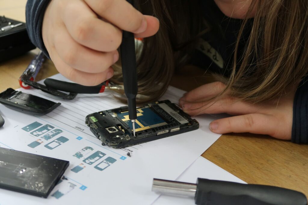 Eine Schülerin löst mit einem sehr feinen Schraubendreher eine kleine Schraube neben der Platine eines geöffneten Handys. Vor ihr liegt einen ausgedruckter technischer Bauplan.
